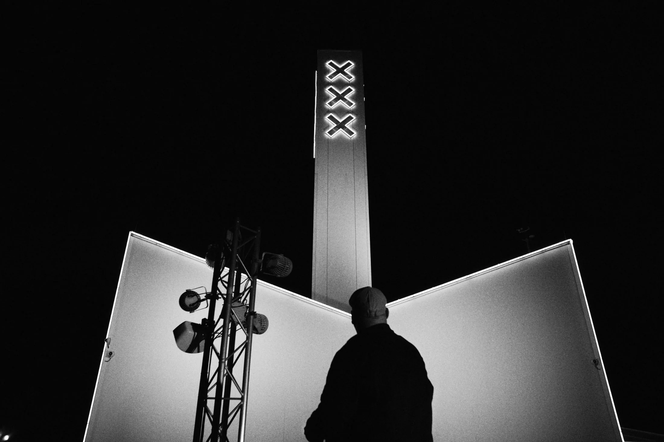 Black-and-white night view of A’DAM Tower in Amsterdam with its three illuminated crosses, a figure in the foreground